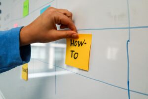 Close-up of a hand placing a yellow 'How-To' sticky note on a whiteboard for planning.